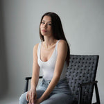 Young woman in a white tank top and jeans sitting on a black chair against a neutral background.