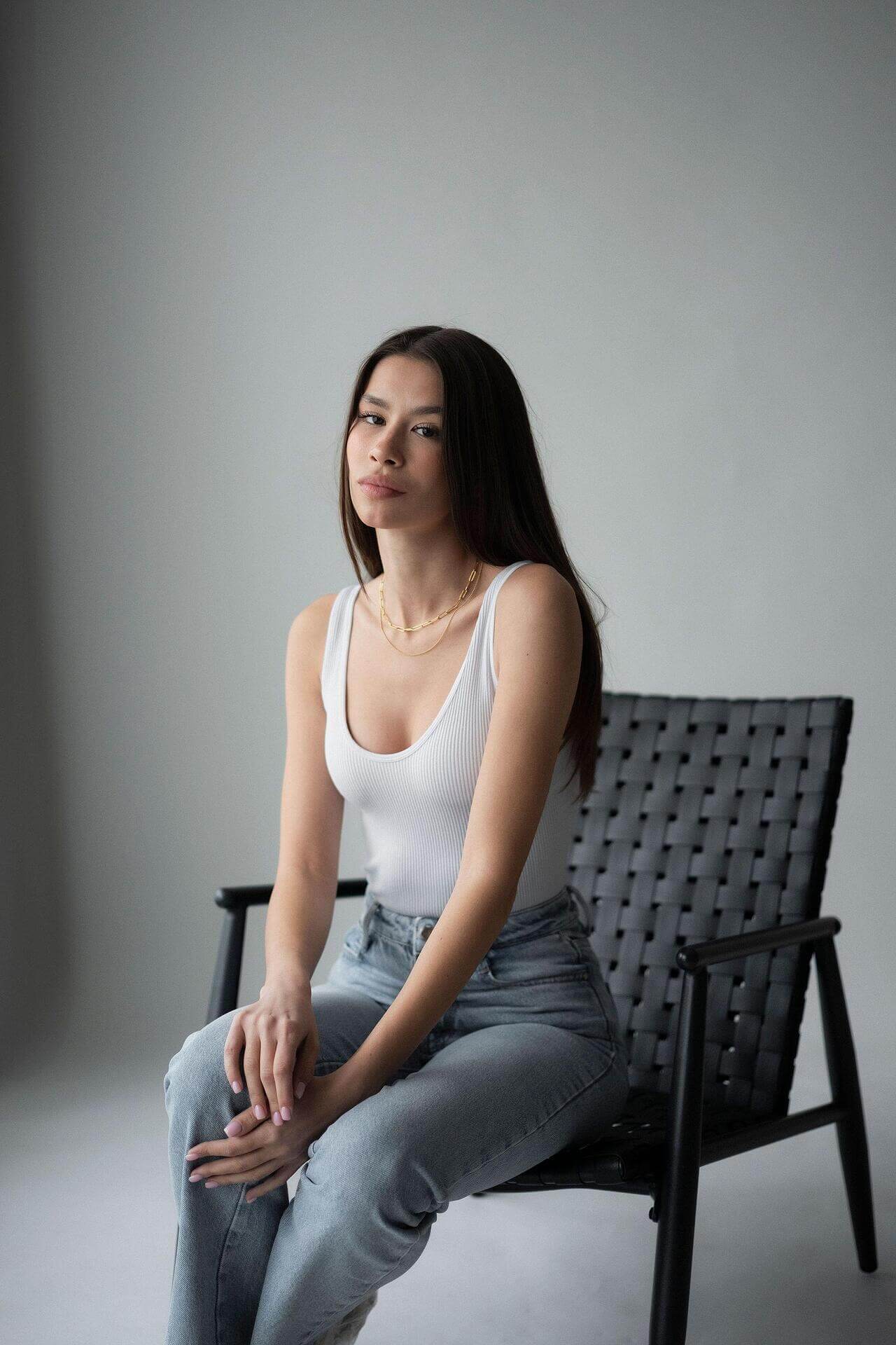 Young woman in a white tank top and jeans sitting on a black chair against a neutral background.