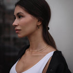 Млада жена с двойно златисто колие и обеци, подчертаваща елегантен стил на фона на неясен градски пейзаж.